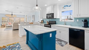 A modern kitchen with white cabinets, a blue accent wall, a large island, and a view of the outdoors. - Hook, Wine & Sinker by Miss Kitty’s (Rockport)
