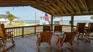 A wooden deck with rocking chairs overlooking a beach, an American flag, and a view of the ocean and buildings. - Hook, Wine & Sinker by Miss Kitty’s (Rockport)