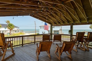 A wooden deck with rocking chairs overlooking a beach, an American flag, and a view of the ocean and buildings.