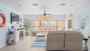 A spacious living room with a beige sofa, a flat-screen TV, a ceiling fan, and a large window with blinds. - Hook, Wine & Sinker by Miss Kitty’s (Rockport)