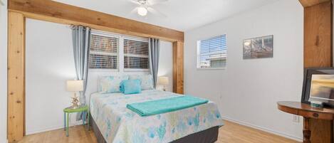 A bedroom with a bed, a ceiling fan, a window with blinds, a small table with a lamp, and a picture frame on the wall.