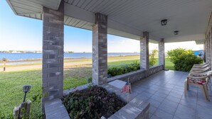 A covered patio with a view of a body of water, stone pillars, a flag, and outdoor furniture. - Attitude Adjustment by Miss Kitty’s (Rockport)