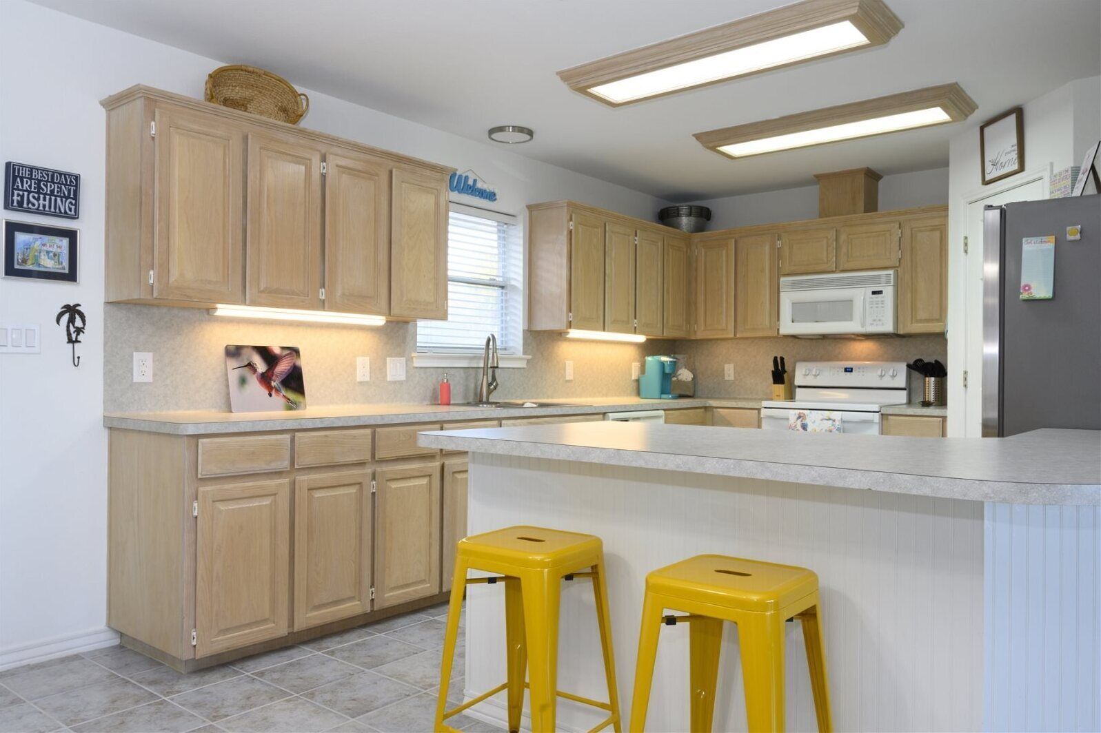 A modern kitchen with wooden cabinets, a central island, yellow stools, a microwave, and a refrigerator.