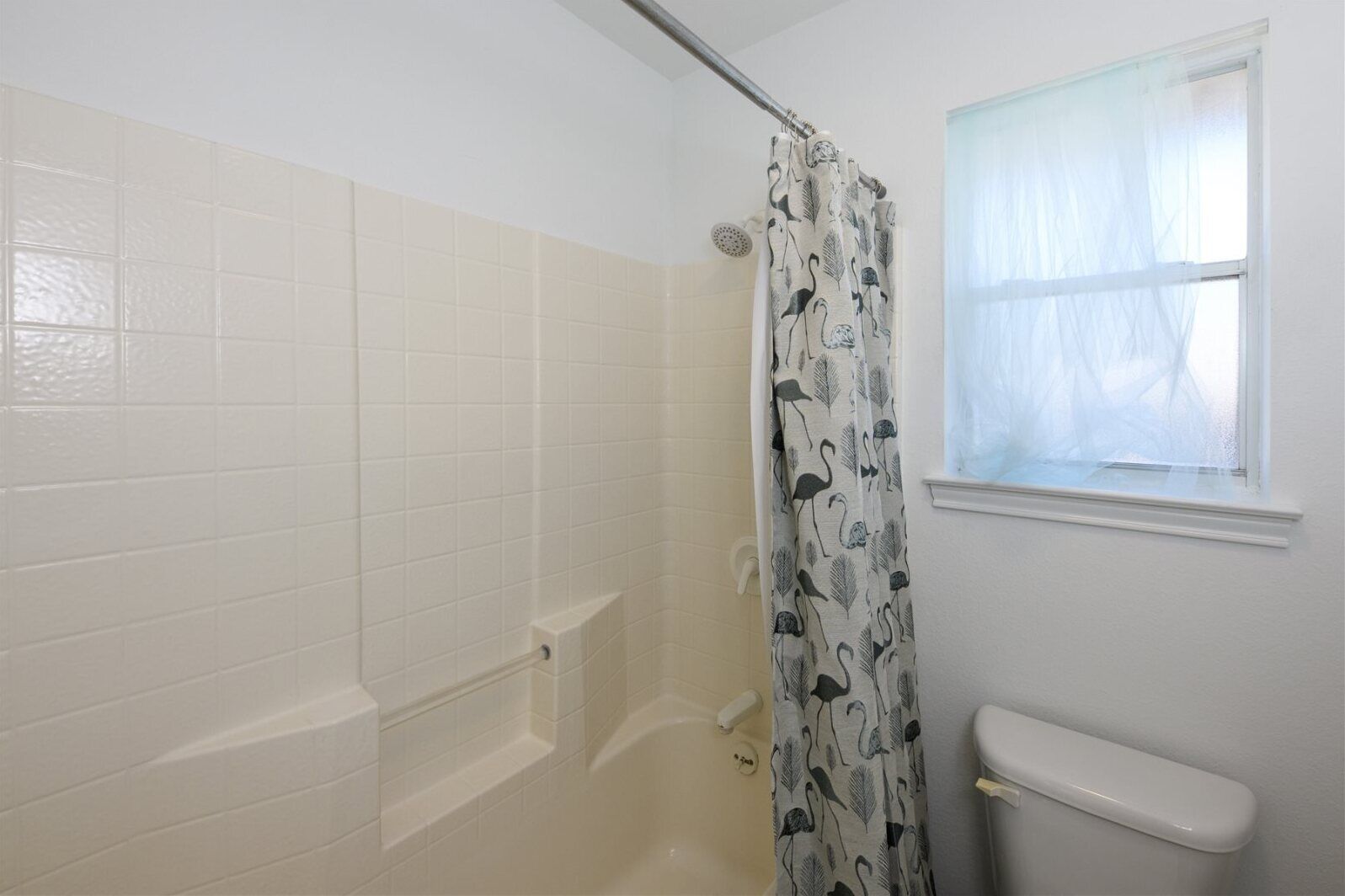 A bathroom with white tiled walls, a shower with a glass door, a toilet, and a window with sheer curtains.
