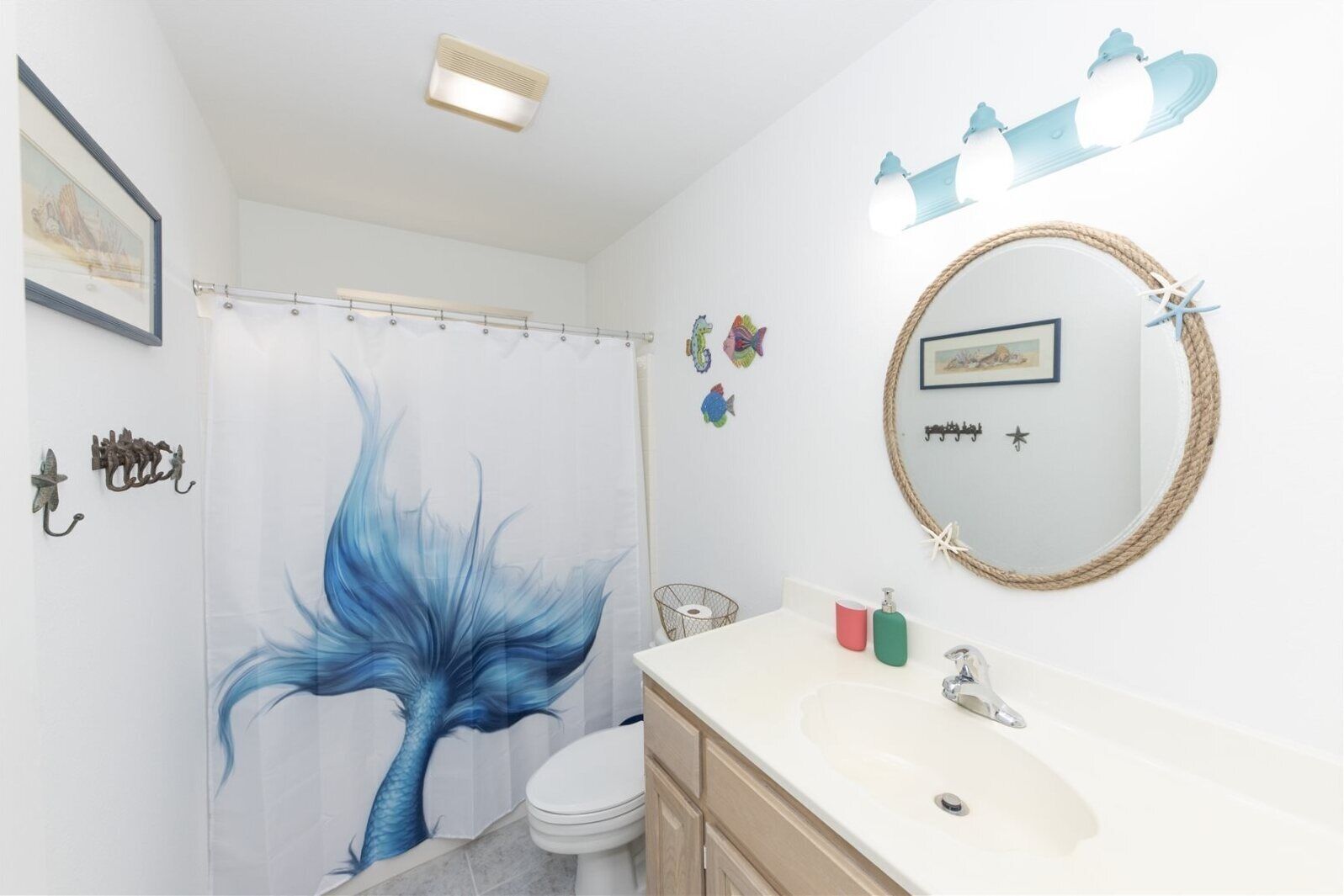 Ein Badezimmer mit Duschvorhang, der ein blaues Meerjungfrauen-Design zeigt, einem weißen Waschbecken mit rundem Spiegel und einer Toilette.