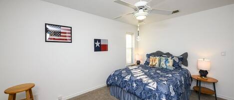 A bedroom with a bed, a ceiling fan, a nightstand with a lamp, and two flags on the wall.