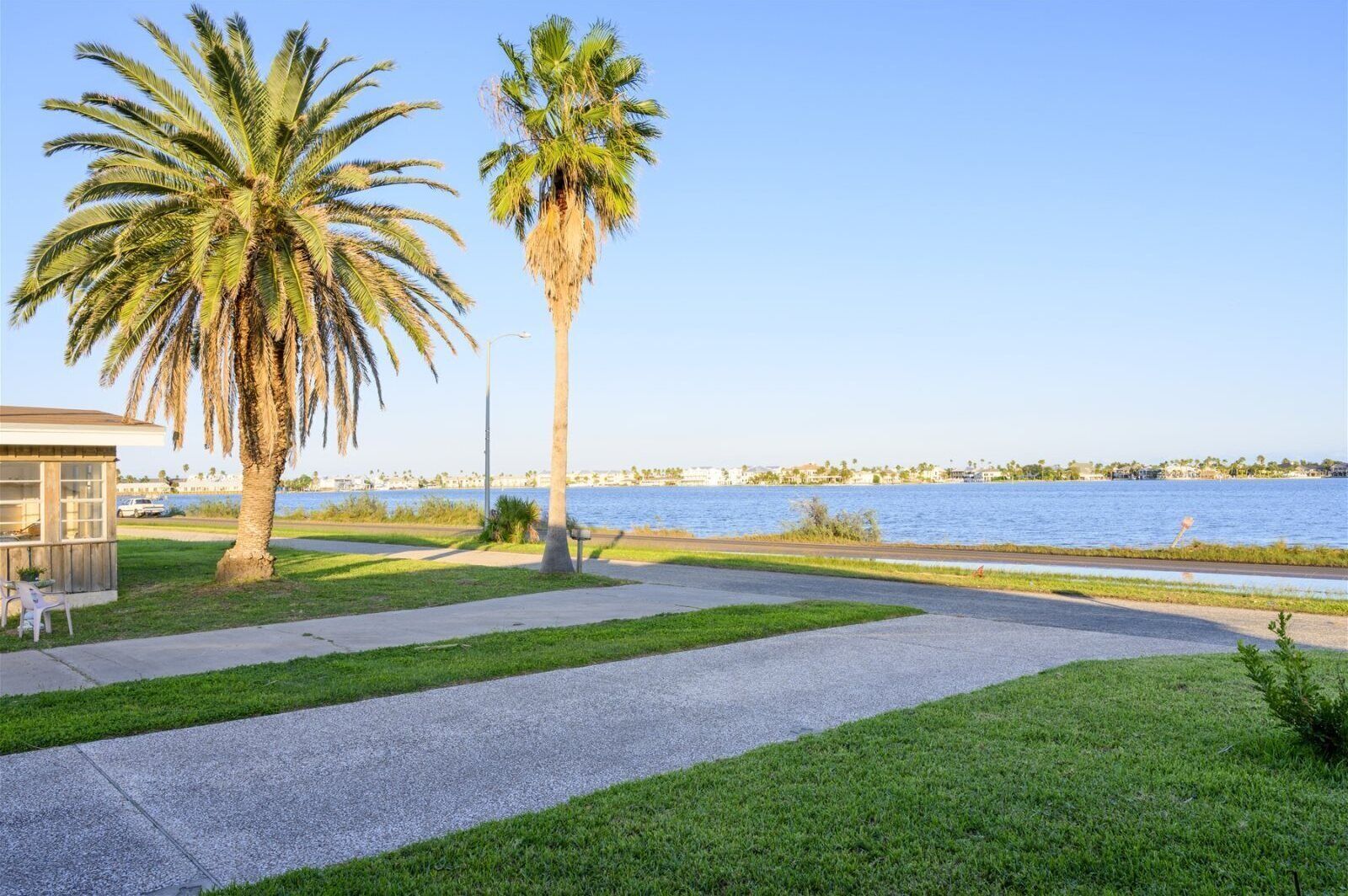 A waterfront park with a paved walkway, palm trees, and a small building with a glass facade.