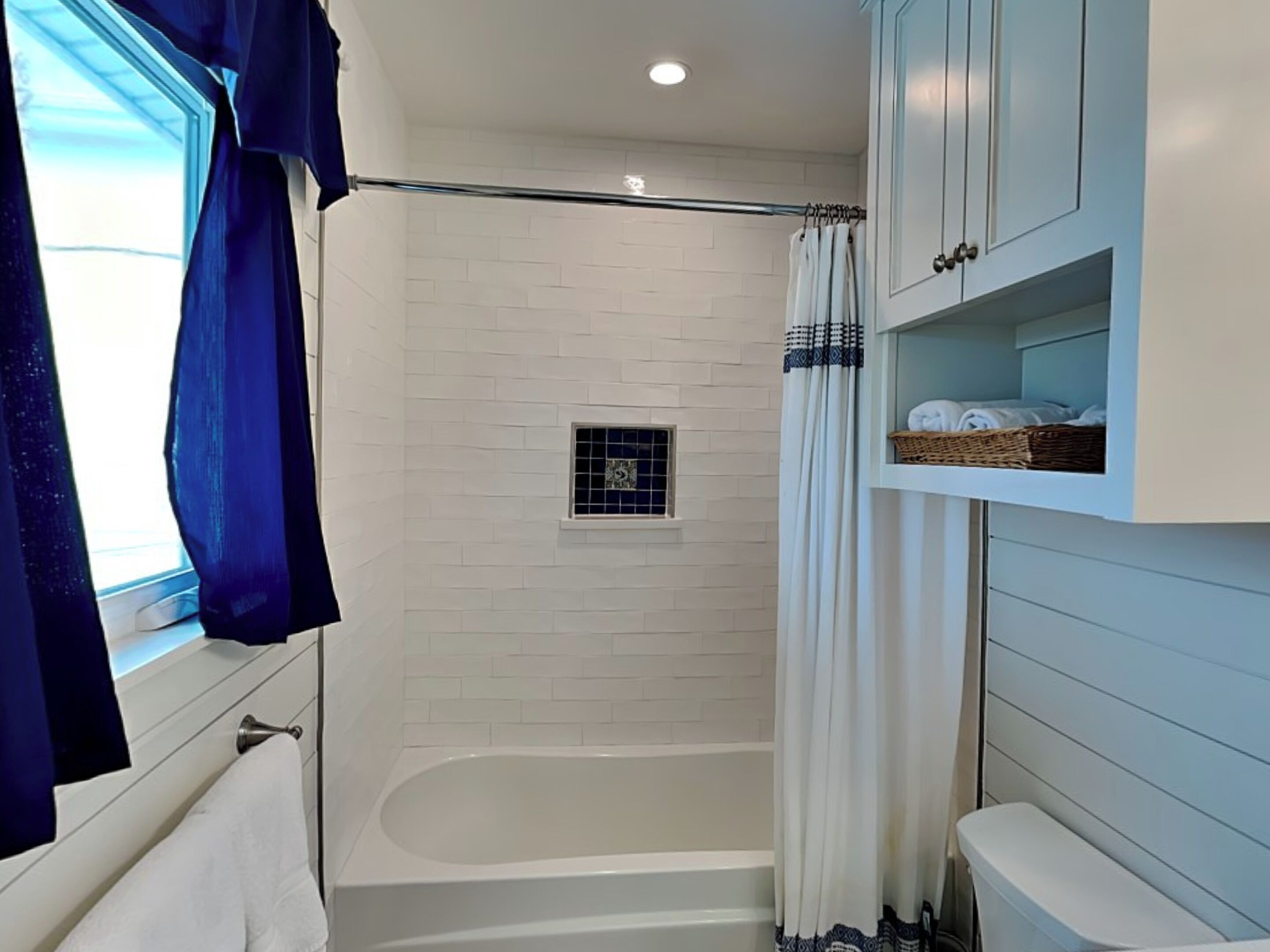 A bathroom with a white bathtub, a shower with a glass door, a window with blue curtains, and a shelf with towels and a basket.
