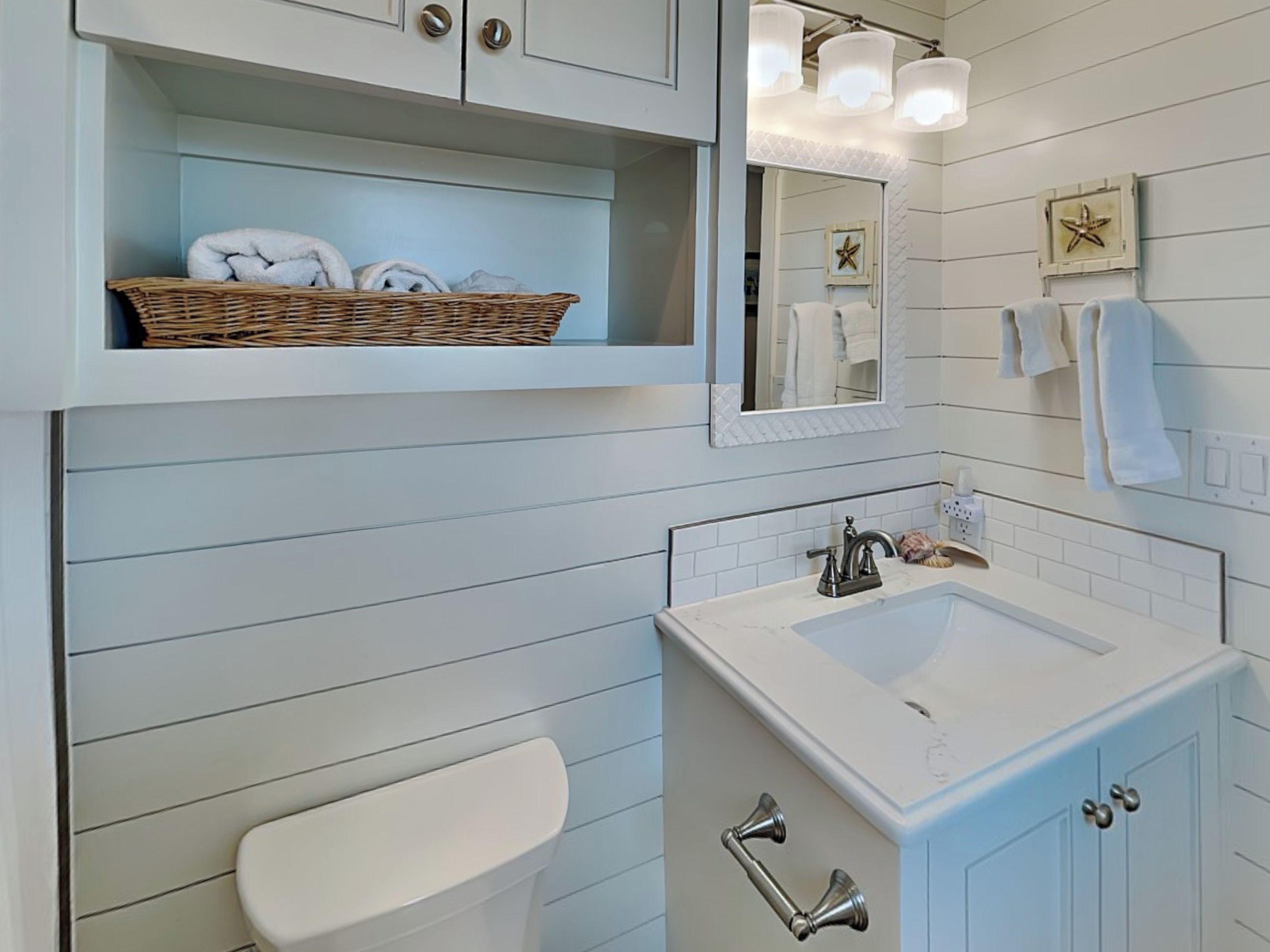 A bathroom with a white sink, a toilet, a wicker basket with towels, and a mirror with lights.