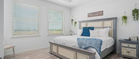 A bedroom with a large bed, a ceiling fan, two windows with blinds, a bedside table, and a nightstand.