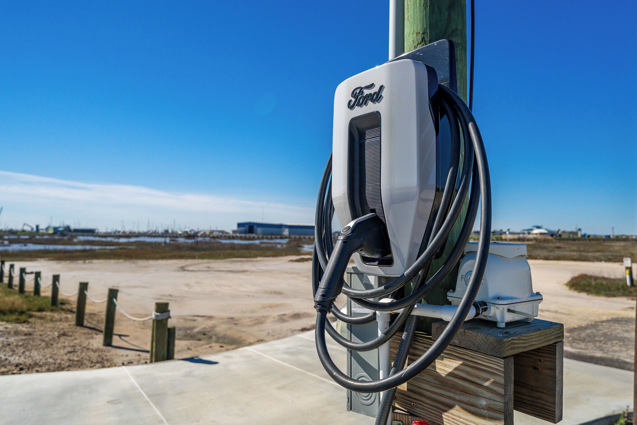 A Ford electric vehicle charging station with a clear view of the surrounding area, including a sandy landscape and distant buildings.