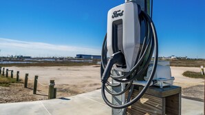 A Ford electric vehicle charging station with a clear view of the surrounding area, including a sandy landscape and distant buildings. - Birds on the Bay by Miss Kitty’s (Rockport)