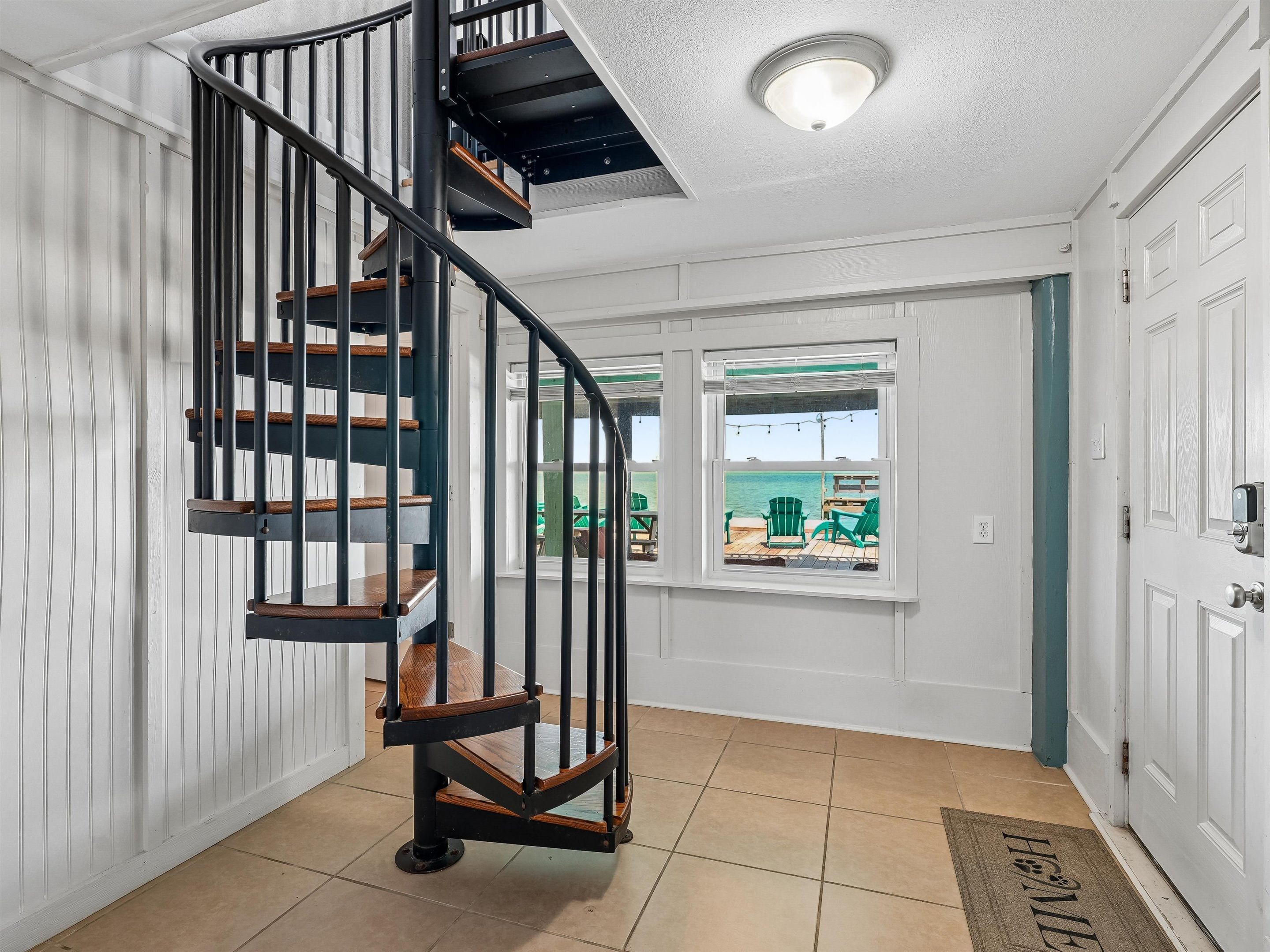 A spiral staircase with wooden steps and black railings, leading to a landing with a view of outdoor seating by a body of water.