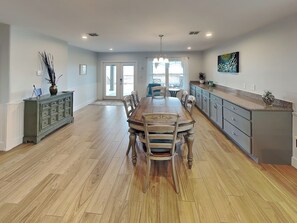 A spacious dining area with a wooden table and chairs, a kitchen with grey cabinets, and a large window allowing natural light. - Trout Tank by Miss Kitty’s (Rockport)