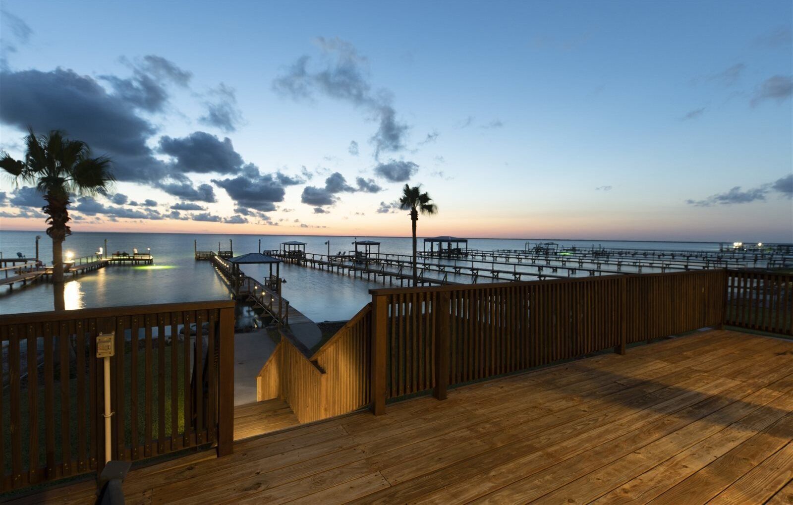 A wooden deck with a view of a pier extending into the water, palm trees, and a sunset sky.