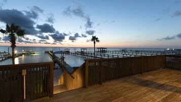 A wooden deck with a view of a pier extending into the water, palm trees, and a sunset sky.