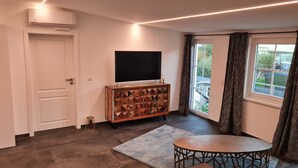 A modern living room with a flat-screen TV, a wooden cabinet with geometric patterns, a round table, and a view of a green landscape through a window with curtains. - Luxurious apartment (new build) with feel-good guarantee.... Temporary living (Ispringen)
