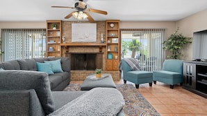 A living room with a fireplace, a ceiling fan, and a large window with blinds. - Gone Coastal by Miss Kitty’s (Rockport)