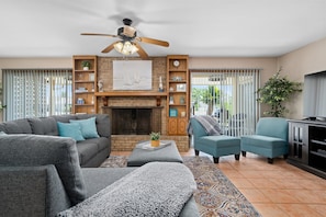 A living room with a fireplace, a ceiling fan, and a large window with blinds. - Gone Coastal by Miss Kitty’s (Rockport)