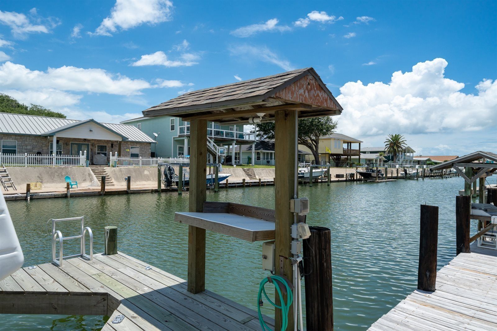 Un muelle de madera con un pequeño refugio, un barco y un puerto deportivo con casas al fondo.