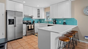 A modern kitchen with white cabinets, stainless steel appliances, a turquoise backsplash, and a bar area with stools. - Gone Coastal by Miss Kitty’s (Rockport)