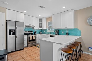 A modern kitchen with white cabinets, stainless steel appliances, a turquoise backsplash, and a bar area with stools. - Gone Coastal by Miss Kitty’s (Rockport)