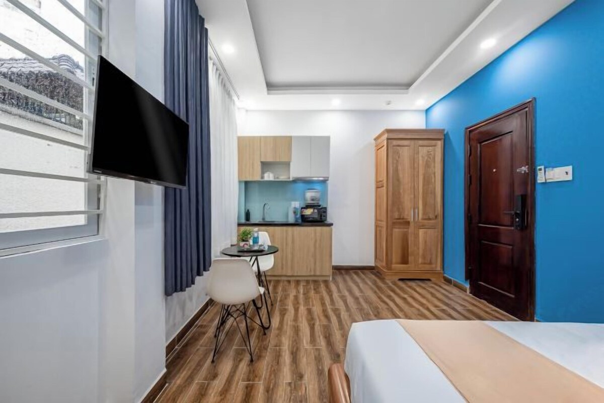 A hotel room with a bed, a TV mounted on the wall, a small kitchen area, and a wooden wardrobe.