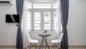 Una habitación con una televisión montada en la pared, una mesa redonda con dos sillas blancas y una ventana con cortinas.