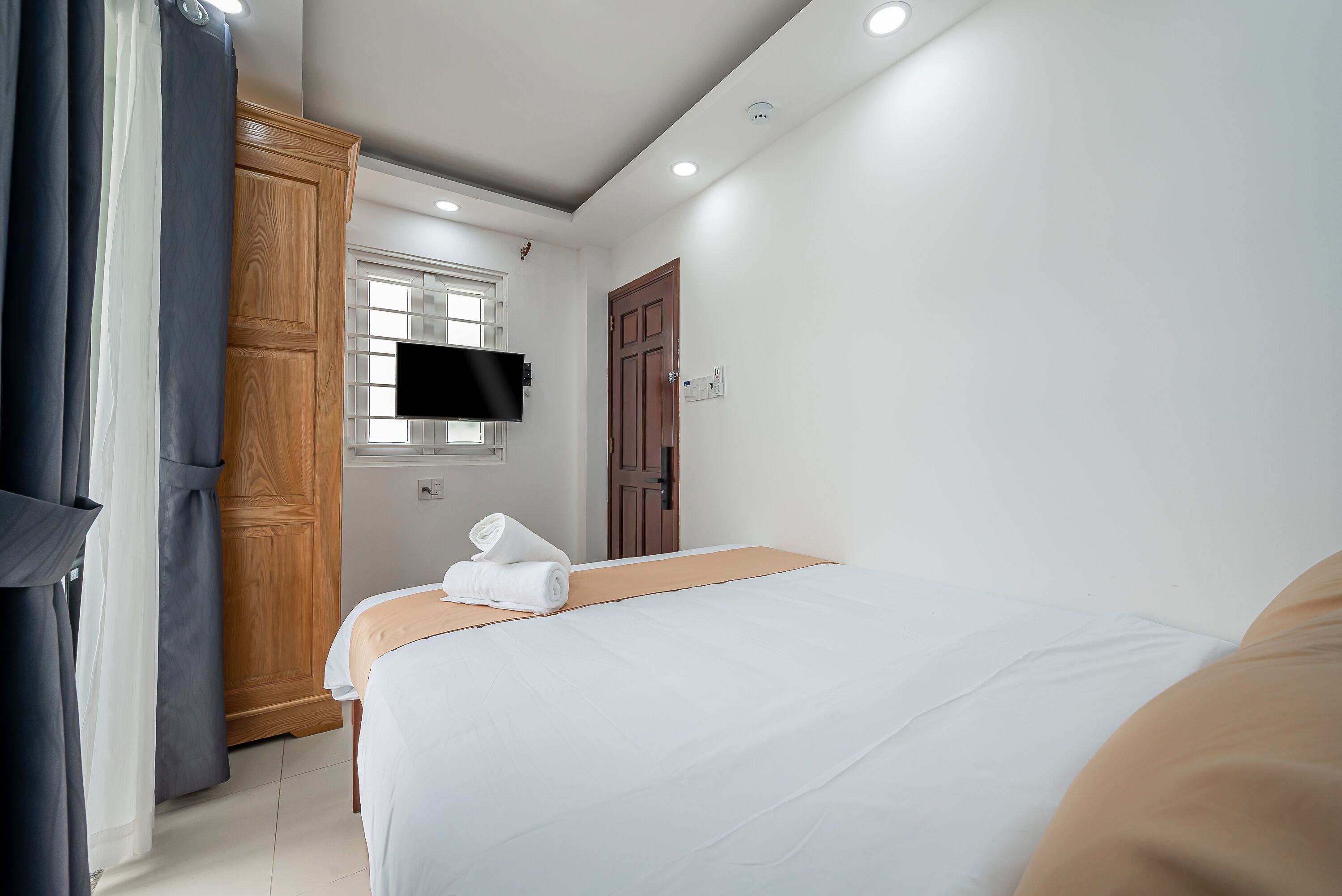 A hotel room with a bed, a wooden wardrobe, a television mounted on the wall, and a window with curtains.