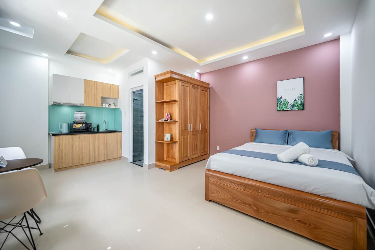 A modern hotel room with a kitchenette, a wooden wardrobe, a bed with a blue and white bedspread, and a framed picture on the wall.