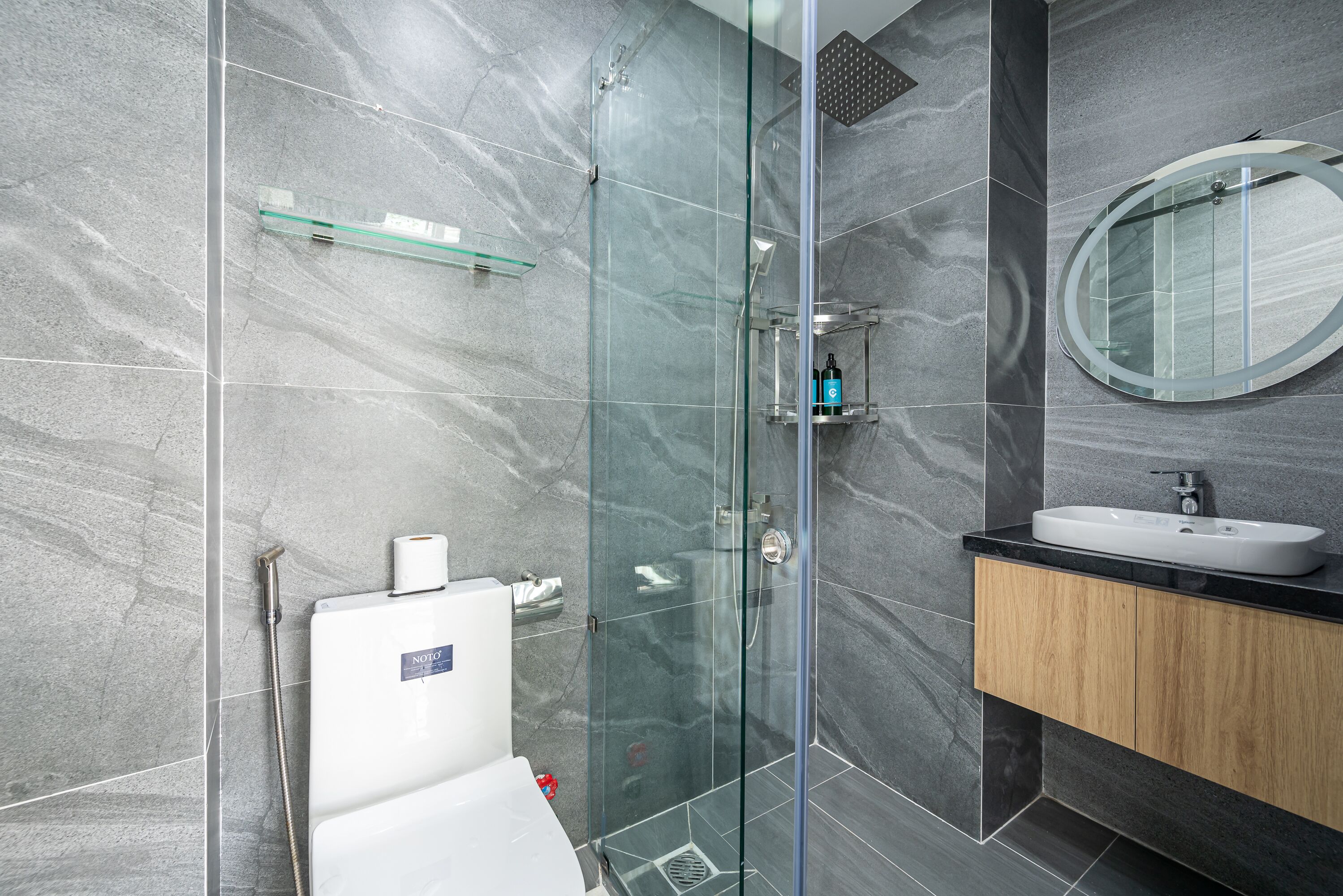 Une salle de bain moderne avec une douche à parois de verre, des toilettes blanches, un lavabo doté d’un plan de travail noir et un meuble en bois.