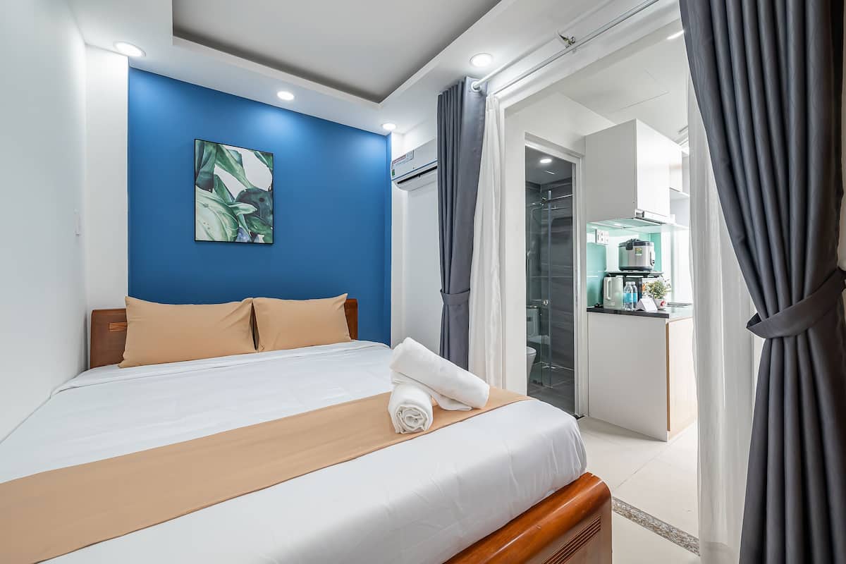 A hotel room with a neatly made bed, a blue accent wall, and a view of the kitchen and bathroom through an open door.