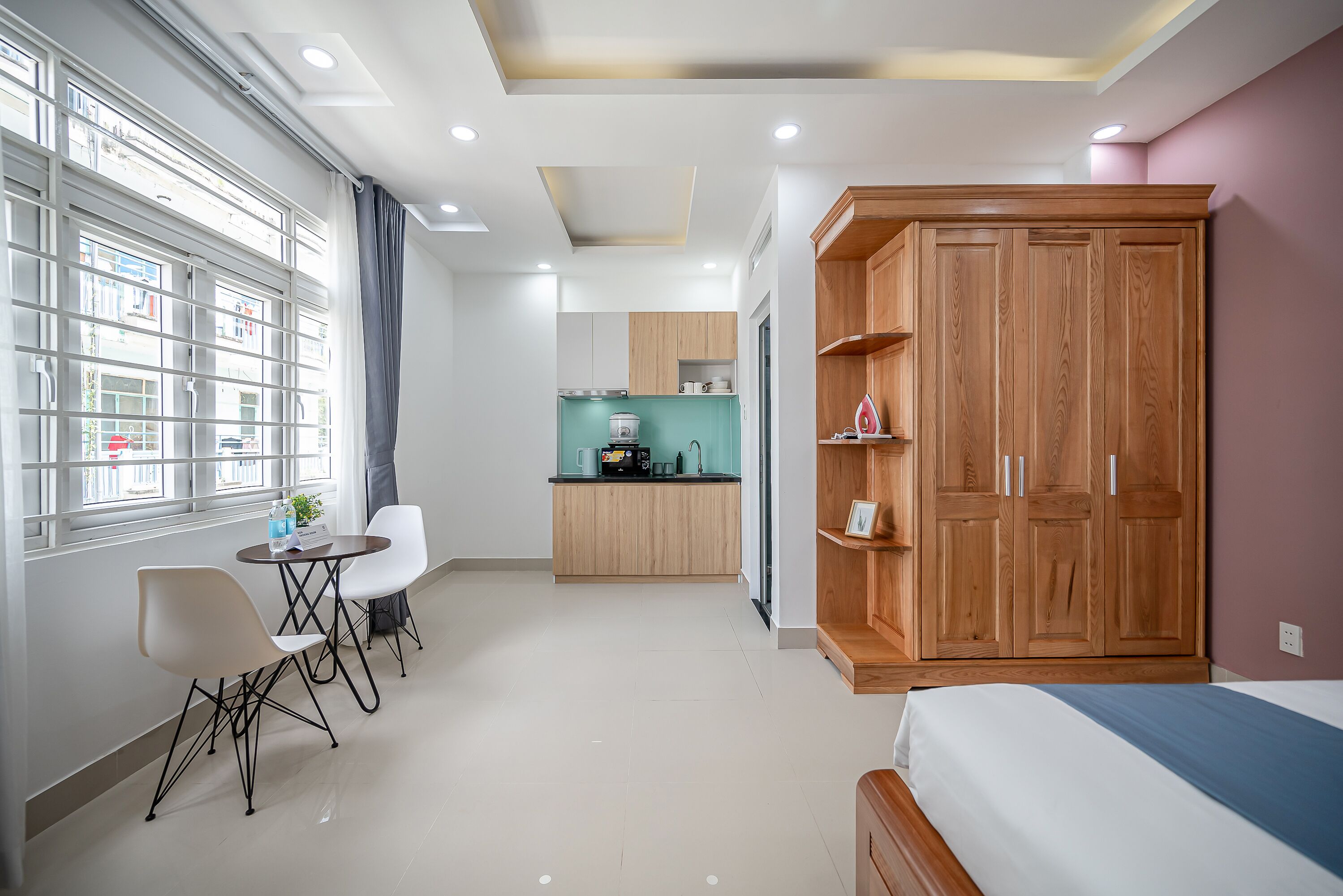 A modern hotel room with a bed, a dining area with chairs, a kitchenette, and a large wardrobe.