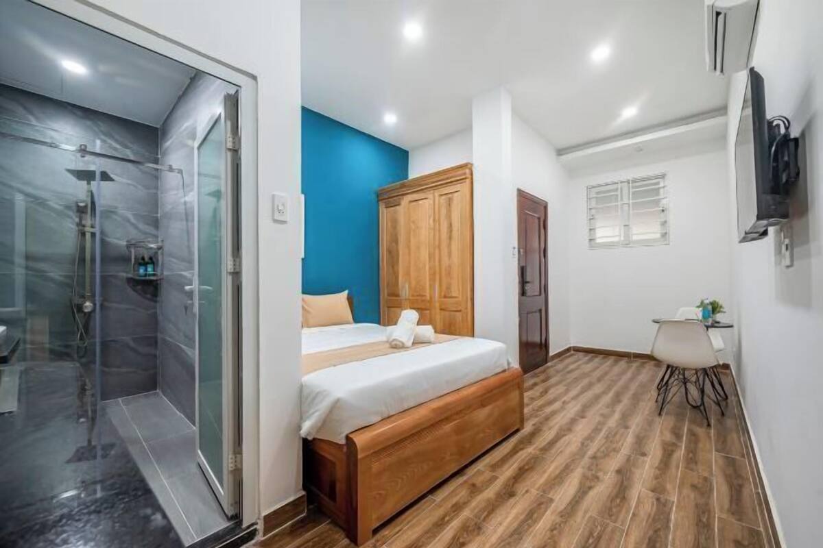 A bedroom with a glass shower, a bed with a wooden headboard, a blue accent wall, and a small table with a chair.
