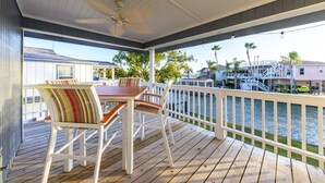 A covered outdoor deck with a table and chairs, a railing, and a view of a marina with boats and houses. - Bayside Blessing by Miss Kitty’s (Rockport)