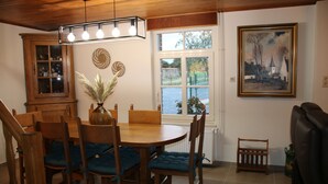 Een eethoek met een houten tafel en stoelen, een kast, een vaas met gedroogde planten en een raam met uitzicht op de omgeving.