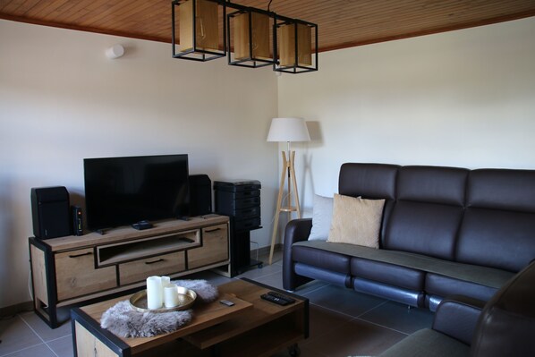 Een moderne woonkamer met een flatscreen televisie, een houten tv-meubel, een leren bank en een bijzettafeltje met een afstandsbediening en kaarsen.