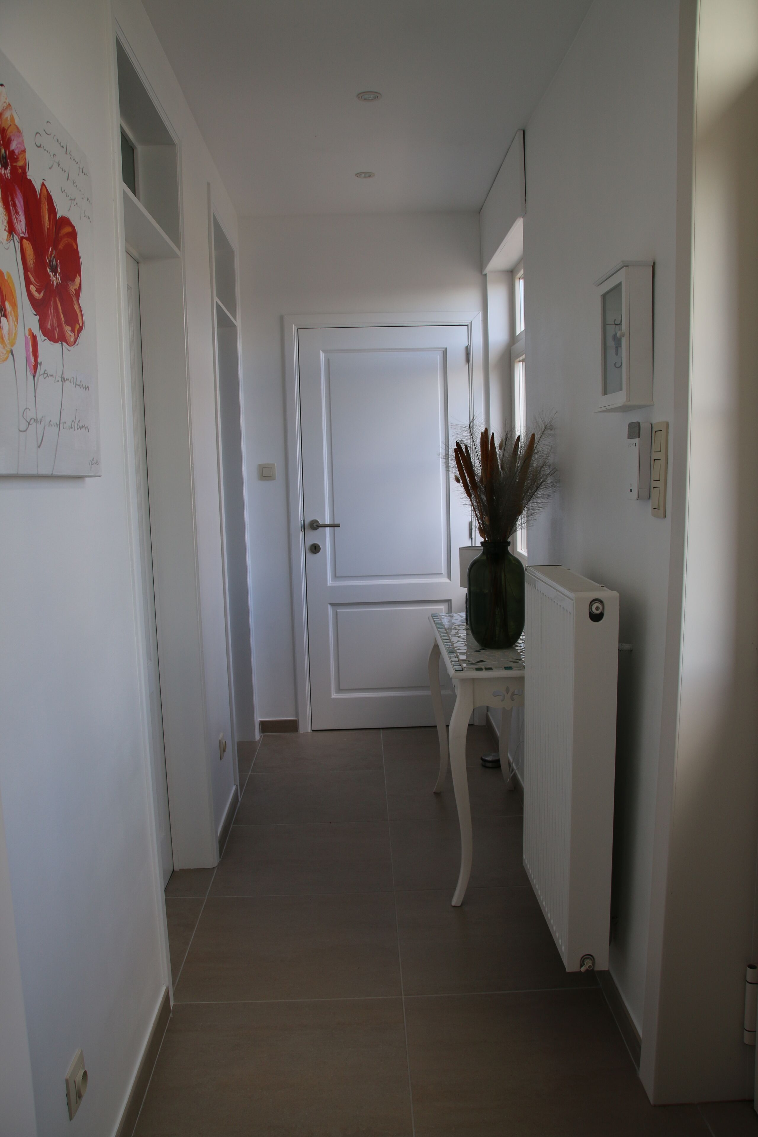 Un couloir avec une porte blanche, un radiateur et une petite table sur laquelle se trouve un vase.