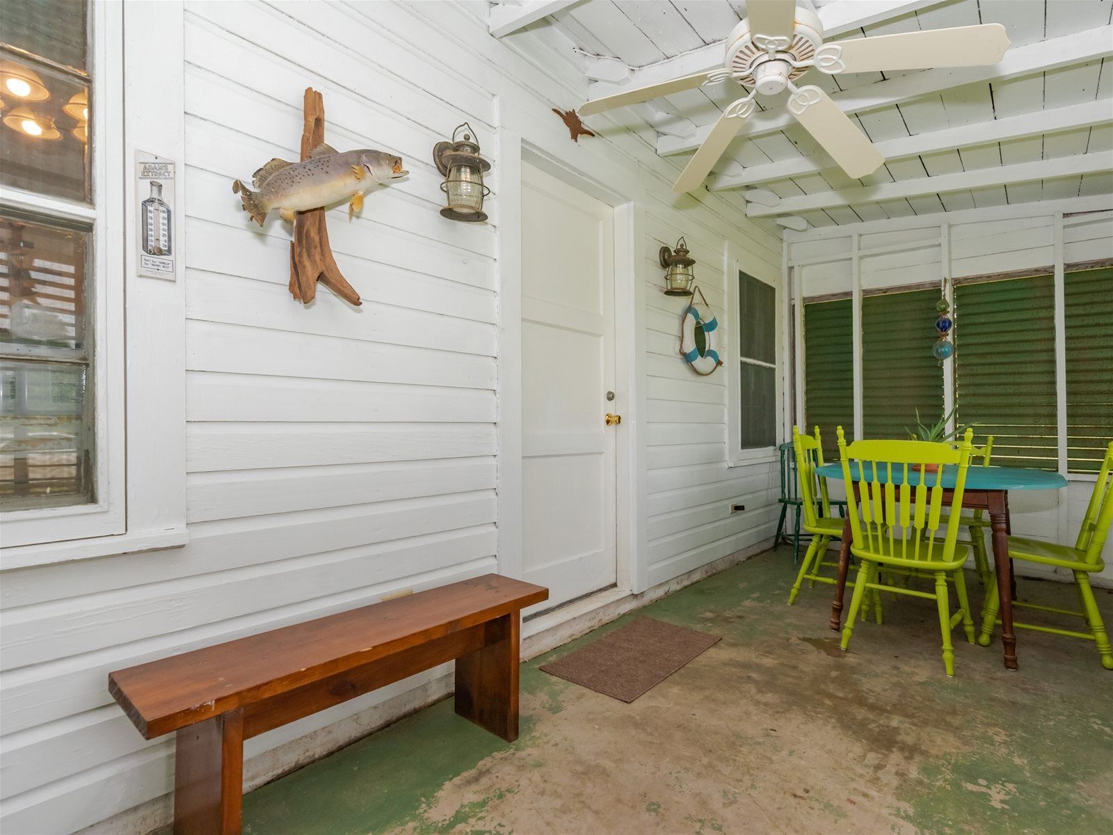 A covered porch with a wooden bench, a table with chairs, a ceiling fan, and decorative items including a fish, a lantern, and a life preserver.
