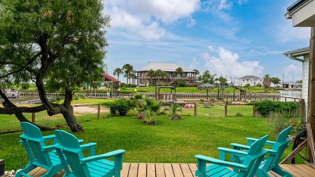 A wooden deck with turquoise chairs, a grassy area with a fence, and a view of a large building across a body of water. - Knot A Care by Miss Kitty’s (Rockport)