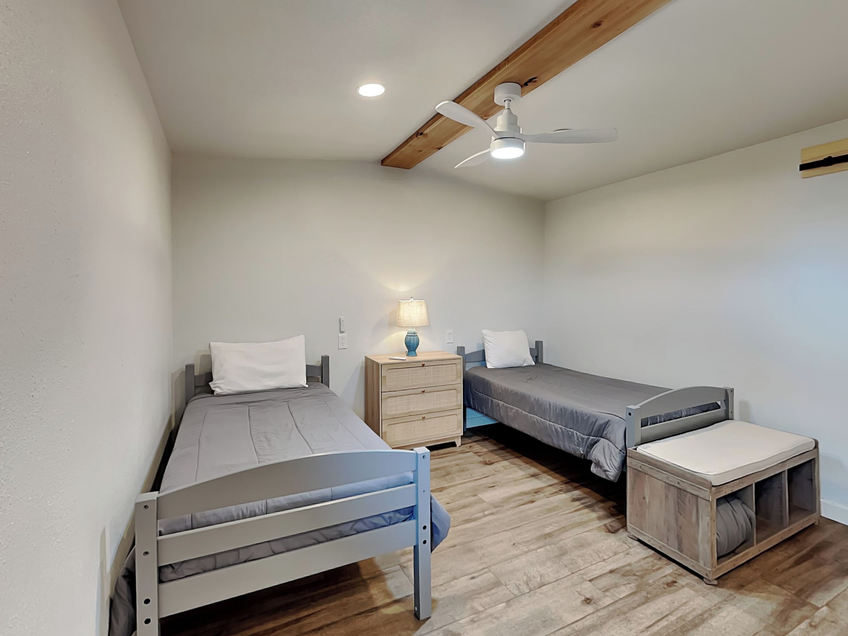 A compact bedroom with two single beds, a nightstand with a lamp, a ceiling fan, and a wooden floor.