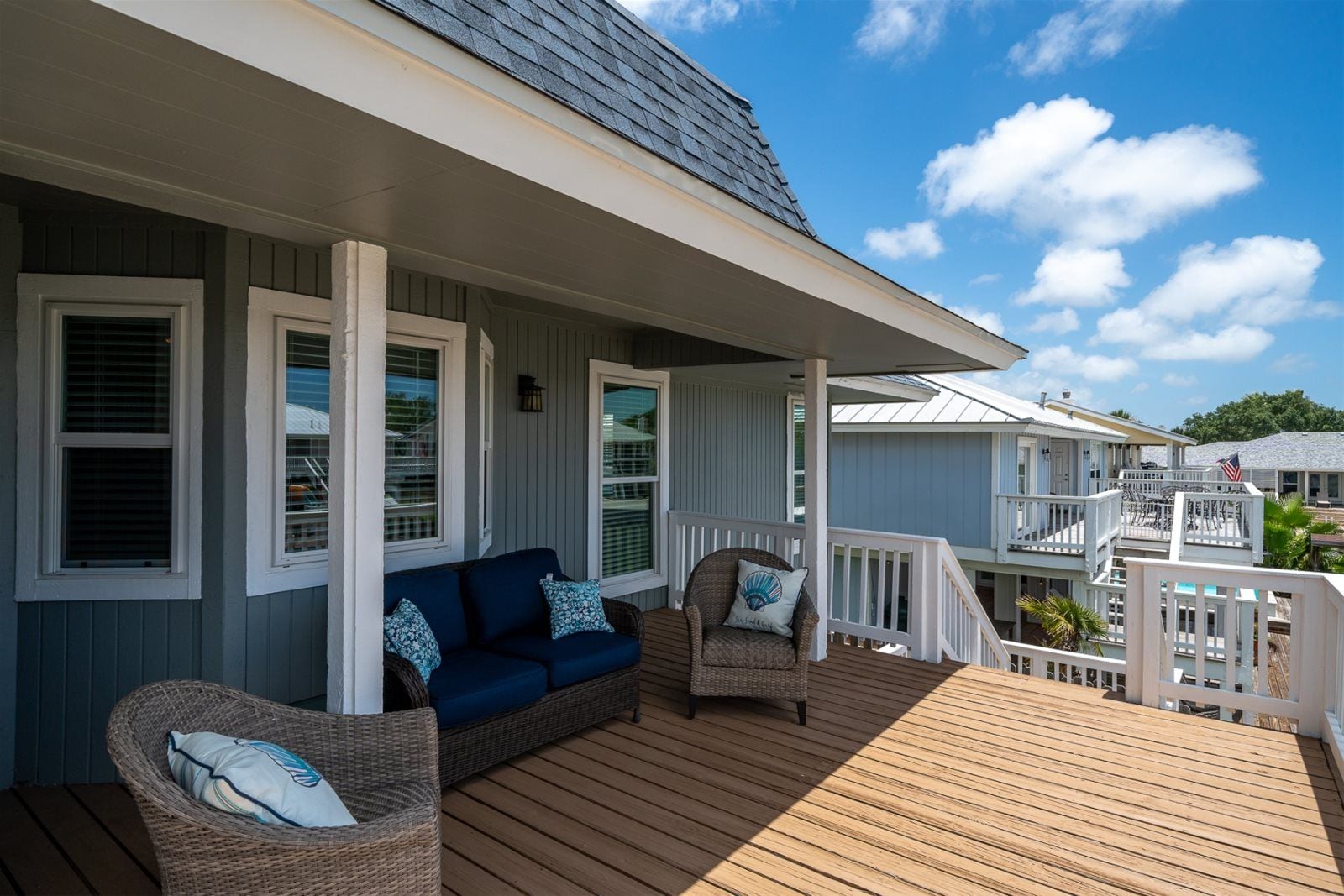 A covered deck with a blue sofa and wicker chairs, wooden railings, and a view of the surrounding area.