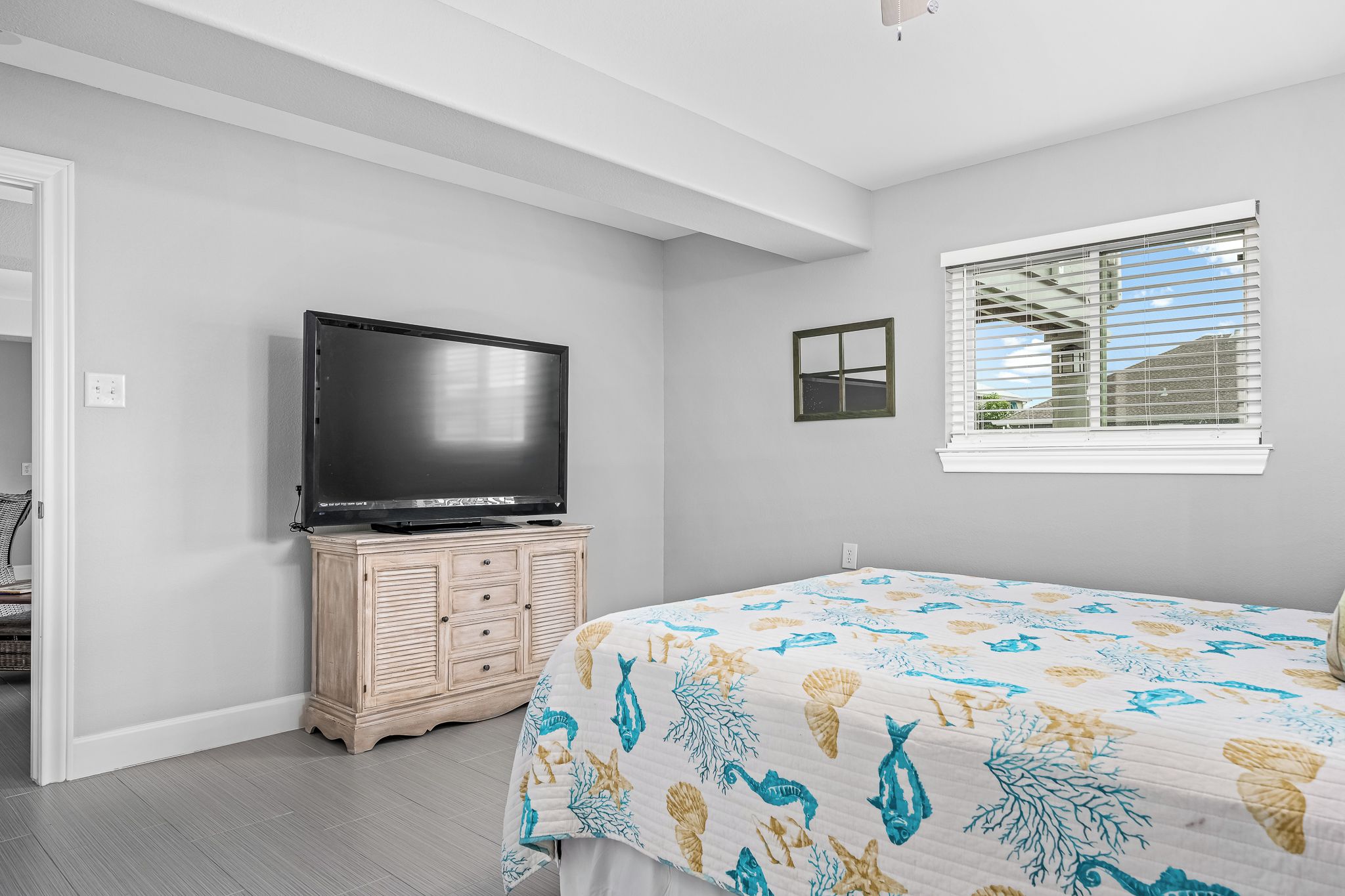 A bedroom with a bed, a TV on a wooden cabinet, a window with blinds, and a door leading to another room.