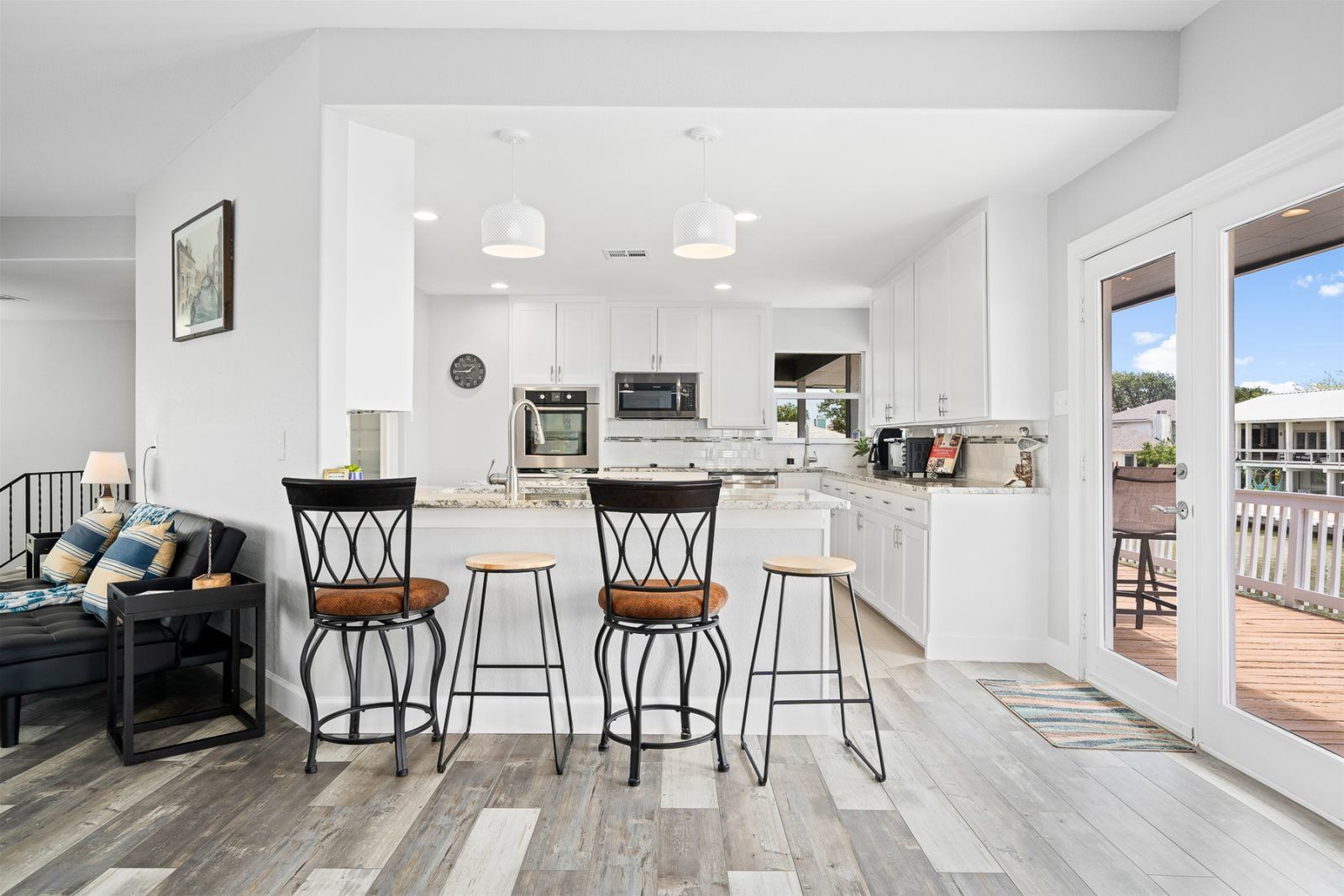 A modern kitchen with white cabinets, a built-in oven, and a microwave. There is a dining area with a black couch, two bar stools, and a small table. The room has wooden flooring and large windows.