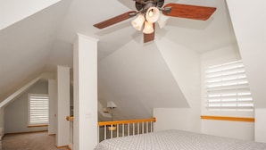Ein Schlafzimmer mit schrägen Wänden, einem Bett mit gemustertem Überwurf, einem Deckenventilator und einem Fenster mit Jalousien.