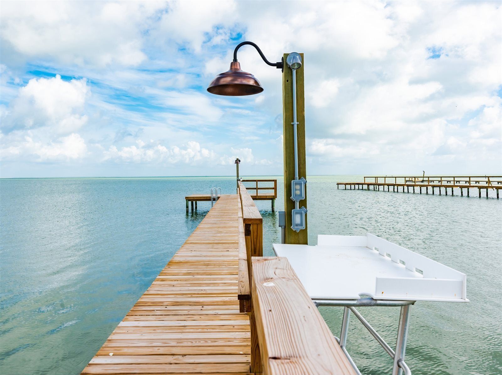 Un muelle de madera que se adentra en aguas tranquilas, con un farol de metal y una silla de lounge blanca.