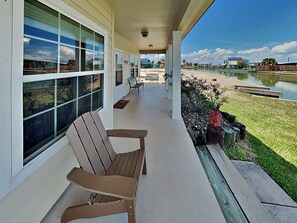 A covered porch with a wooden chair, a bench, and a view of a lake and buildings in the distance. - Mermaid Bay by Miss Kitty’s (Rockport)