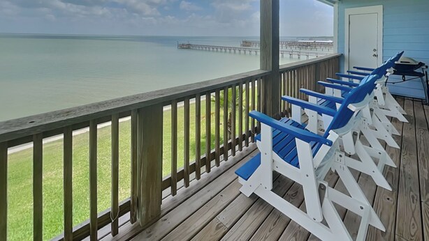 A wooden deck with blue and white Adirondack chairs overlooking a calm sea and a pier. - Copano Delight by Miss Kitty’s (Rockport)