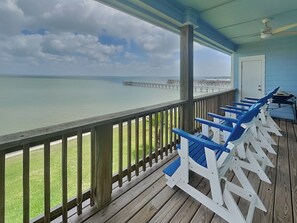 A wooden deck with blue and white Adirondack chairs overlooking a calm sea and a pier. - Copano Delight by Miss Kitty’s (Rockport)