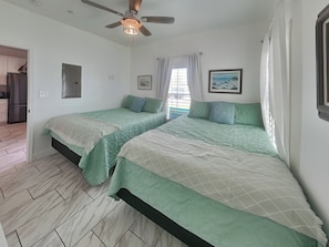 Un dormitorio con dos camas, ventilador de techo, ventana con cortinas y una cocina visible a través de una puerta abierta.
